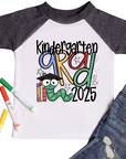 Children's graduation shirt that says 'First Grade Grad 2025' with colorful letters, a cartoon bookworm wearing a graduation cap, and school-themed accessories. Shirt shown with markers and denim jeans.