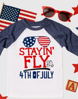 Kids raglan t-shirt with "Stayin’ Fly on the 4th of July" design featuring American flag sunglasses in red, white, and blue
