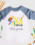Children's shirt with 'fifth grade' and construction-themed design, surrounded by school supplies on a white background.