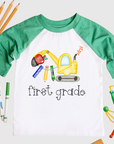 Green and white raglan T-shirt with a watercolor-style construction excavator made from school supplies like a pencil, ruler, and crayon, with the words “first grade” printed below. Surrounded by school-themed props on a white background.