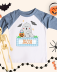 Boys personalized Halloween ghost shirt on a kids 3/4-sleeve raglan with heather blue sleeves and white body. Ghost with pumpkins and blue gingham name box reads “JACOB,” styled with bat cutout, pumpkin bucket, black bead garland, spider, and orange streamers on a cream background.