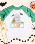 Boys personalized Halloween ghost shirt on a kids 3/4-sleeve raglan with heather green sleeves and white body. Ghost with pumpkins and blue gingham name box reads “OLIVER,” styled with bat cutout, pumpkin bucket, black bead garland, spider, and orange streamers on a cream background.