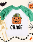 Personalized boys pumpkin shirt on a kids 3/4-sleeve raglan with heather green sleeves and white body. Jack-o’-lantern wearing a cap above the name “CHASE.” Styled flat lay with bat cutout, pumpkin pail, lollipop, spider, skeleton, and black bead garland on a cream background. Kids Halloween name tee, sizes 6M–Youth Large.
