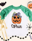 Personalized girls pumpkin shirt on a kids 3/4-sleeve raglan with heather green sleeves and white body. Jack-o’-lantern with a big bow above the name “Sophia.” Flat-lay styling with bat cutout, pumpkin bucket, spider, skeleton, bead garland, and streamers on a cream background. Kids Halloween name tee, sizes 6M–Youth Large.