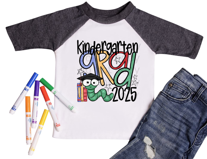 Children's graduation shirt that says 'First Grade Grad 2025' with colorful letters, a cartoon bookworm wearing a graduation cap, and school-themed accessories. Shirt shown with markers and denim jeans.