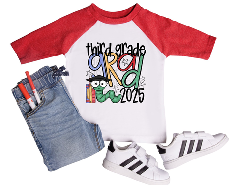 Children's graduation shirt that says 'First Grade Grad 2025' with colorful letters, a cartoon bookworm wearing a graduation cap, and school-themed accessories. Shirt shown with markers, denim jeans, and sneakers.