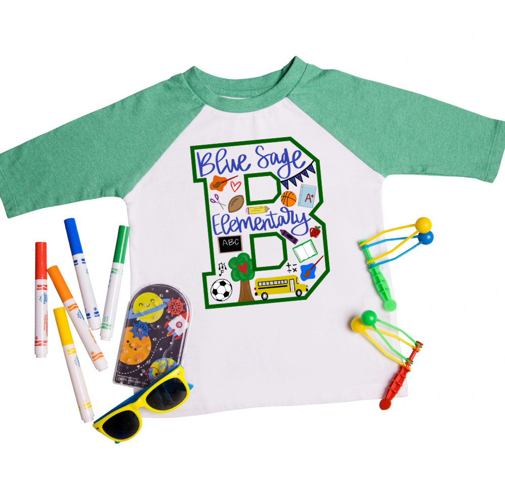 Green-sleeve kids raglan tee with a playful school-themed “B” design for Blue Sage Elementary, decorated with colorful doodles like books, a school bus, sports balls, trees, and pencils. Surrounded by vibrant school supplies, sunglasses, and toys for a fun flat lay presentation.