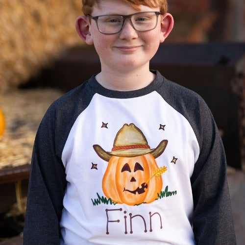 Boy wearing a personalized Halloween shirt with dark gray raglan sleeves and white body. The design features a smiling cowboy jack-o-lantern wearing a tan hat and holding a wheat stem, personalized with the name “Finn.” Perfect for fall photos, pumpkin-patch outings, or Halloween. Soft 65% polyester / 35% cotton blend kids raglan.