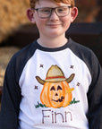 Boy wearing a personalized Halloween shirt with dark gray raglan sleeves and white body. The design features a smiling cowboy jack-o-lantern wearing a tan hat and holding a wheat stem, personalized with the name “Finn.” Perfect for fall photos, pumpkin-patch outings, or Halloween. Soft 65% polyester / 35% cotton blend kids raglan.