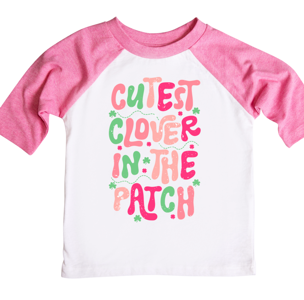 A kids’ St. Patrick’s Day shirt with pink raglan sleeves and a playful design featuring the phrase “Cutest Clover in the Patch” in fun, colorful lettering with green shamrock accents. The shirt is perfect for festive holiday outfits, adding a cheerful touch of Irish charm.