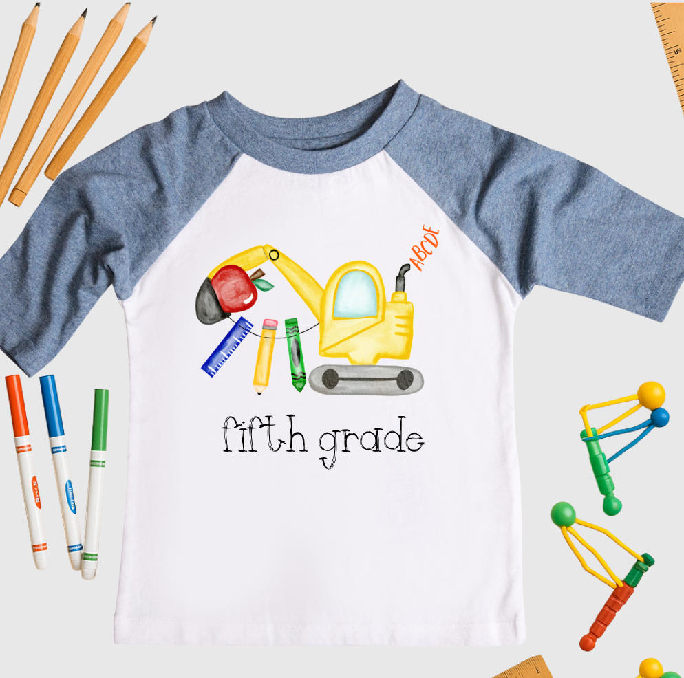 Children's shirt with 'fifth grade' and construction-themed design, surrounded by school supplies on a white background.