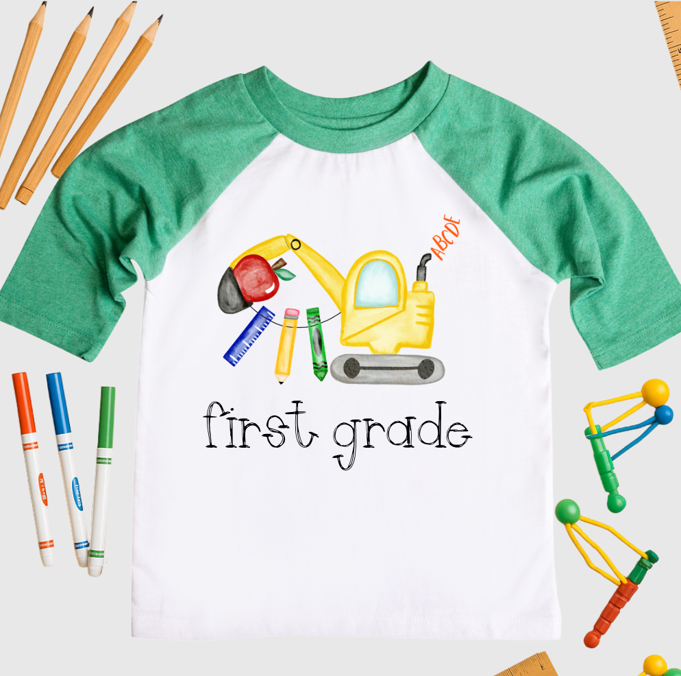 Green and white raglan T-shirt with a watercolor-style construction excavator made from school supplies like a pencil, ruler, and crayon, with the words “first grade” printed below. Surrounded by school-themed props on a white background.