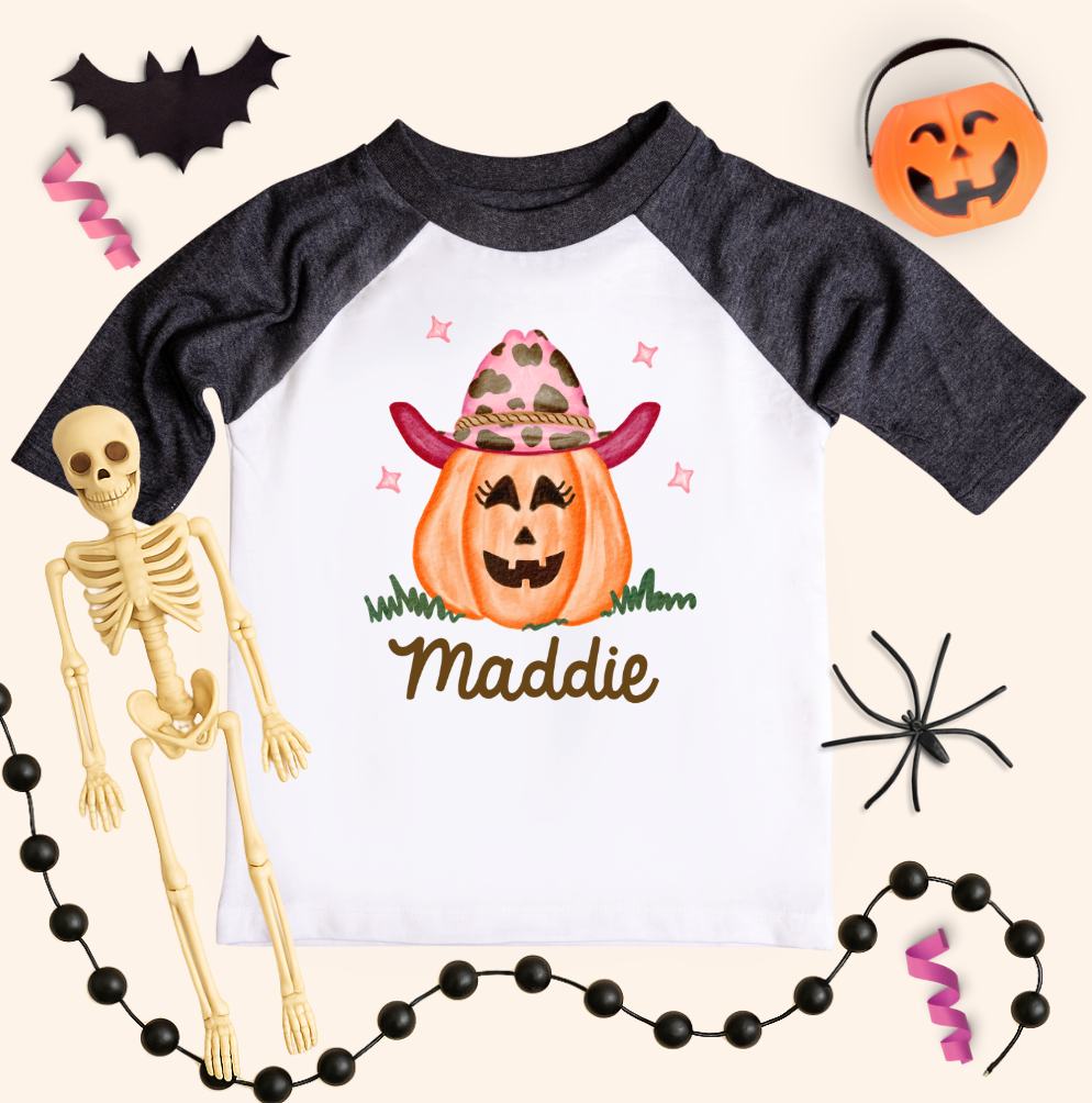 Halloween-themed children&#39;s shirt with a pumpkin design and name &#39;Maddie&#39;, surrounded by Halloween decorations.