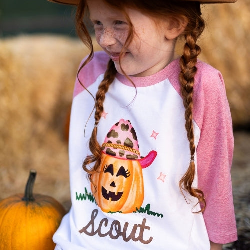 Little girl wearing a personalized pink raglan Halloween shirt featuring a cowgirl jack-o-lantern with a leopard hat and pink brim. The shirt is customized with the name “Scout” in brown lettering. She’s smiling and sitting near hay bales and a pumpkin — perfect for fall photos, pumpkin patch outings, or Halloween. Soft polyester-cotton kids raglan tee.