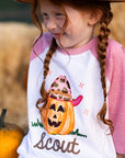 Little girl wearing a personalized pink raglan Halloween shirt featuring a cowgirl jack-o-lantern with a leopard hat and pink brim. The shirt is customized with the name “Scout” in brown lettering. She’s smiling and sitting near hay bales and a pumpkin — perfect for fall photos, pumpkin patch outings, or Halloween. Soft polyester-cotton kids raglan tee.
