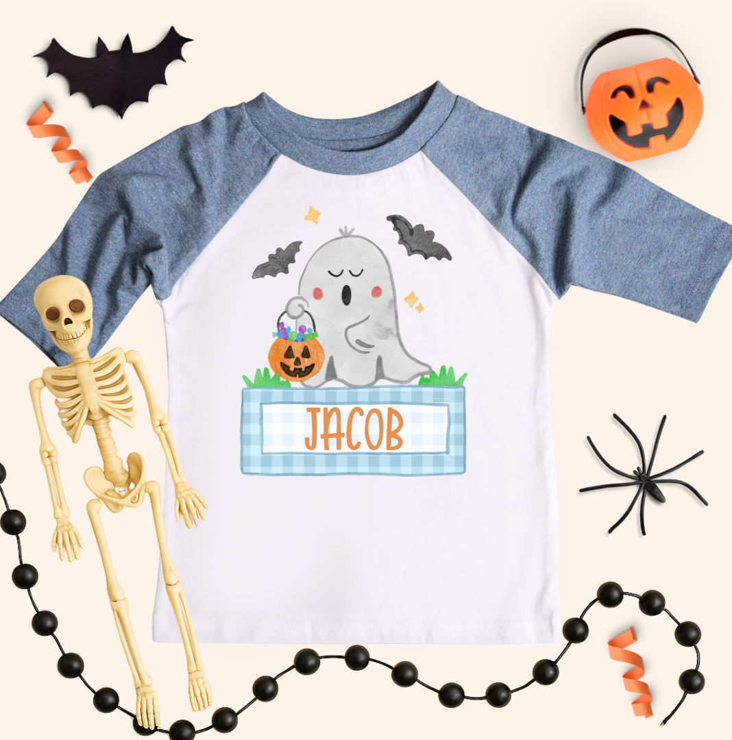 Boys personalized Halloween ghost shirt on a kids 3/4-sleeve raglan with heather blue sleeves and white body. Ghost with pumpkins and blue gingham name box reads “JACOB,” styled with bat cutout, pumpkin bucket, black bead garland, spider, and orange streamers on a cream background.