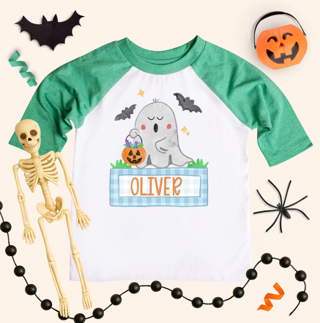 Boys personalized Halloween ghost shirt on a kids 3/4-sleeve raglan with heather green sleeves and white body. Ghost with pumpkins and blue gingham name box reads “OLIVER,” styled with bat cutout, pumpkin bucket, black bead garland, spider, and orange streamers on a cream background.