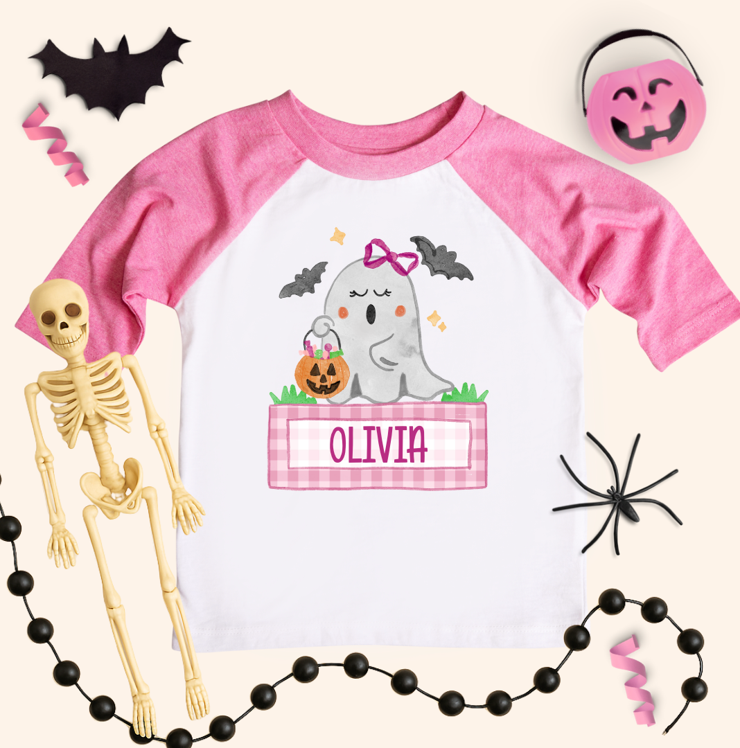Pink and white t-shirt with ghost design and 'Olivia' text, surrounded by Halloween decorations.
