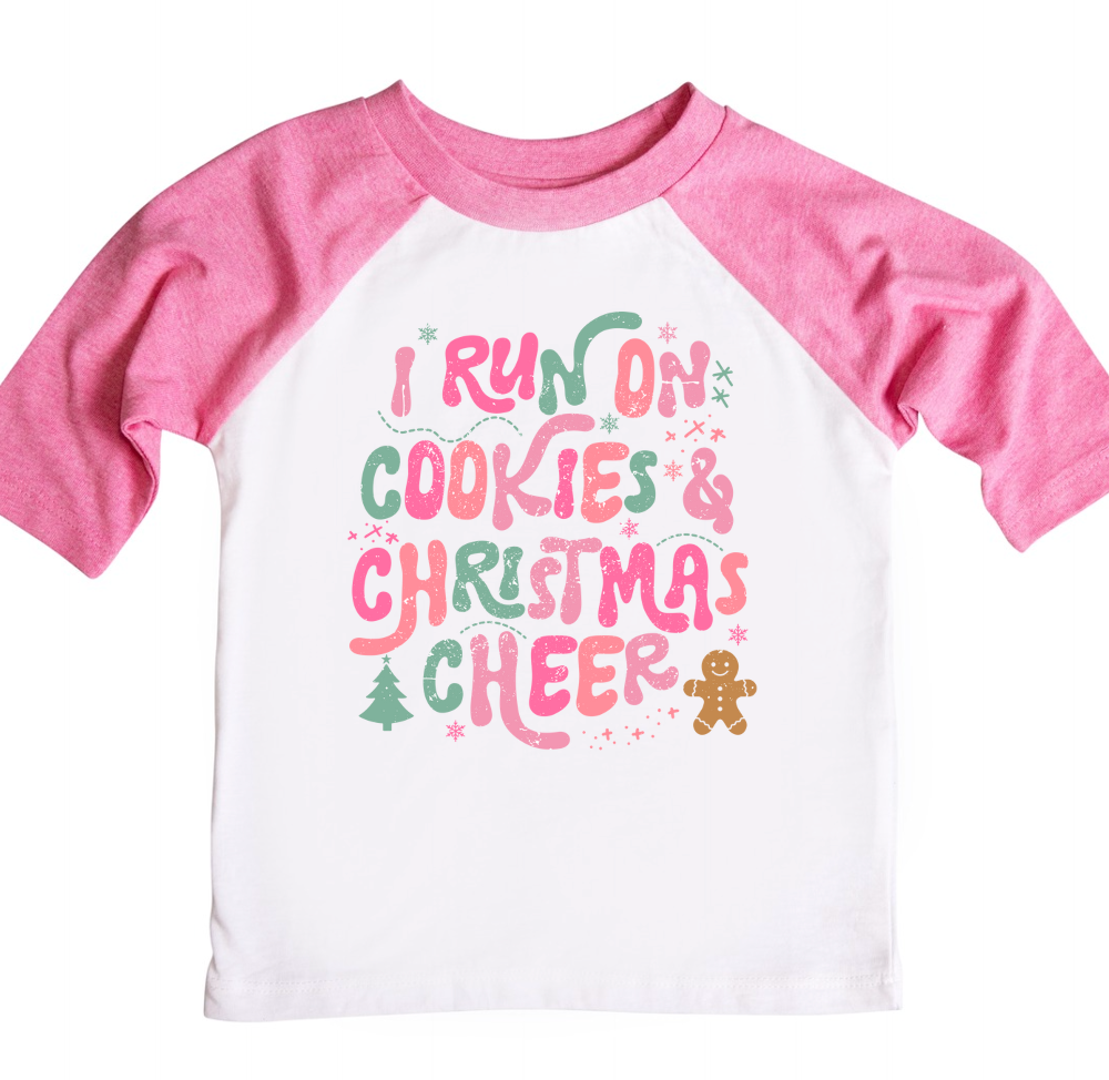Pink and white raglan shirt featuring the festive phrase 'I run on cookies & Christmas cheer' in playful pink, green, and red lettering, surrounded by holiday-themed elements like stars, a Christmas tree, and a gingerbread man.