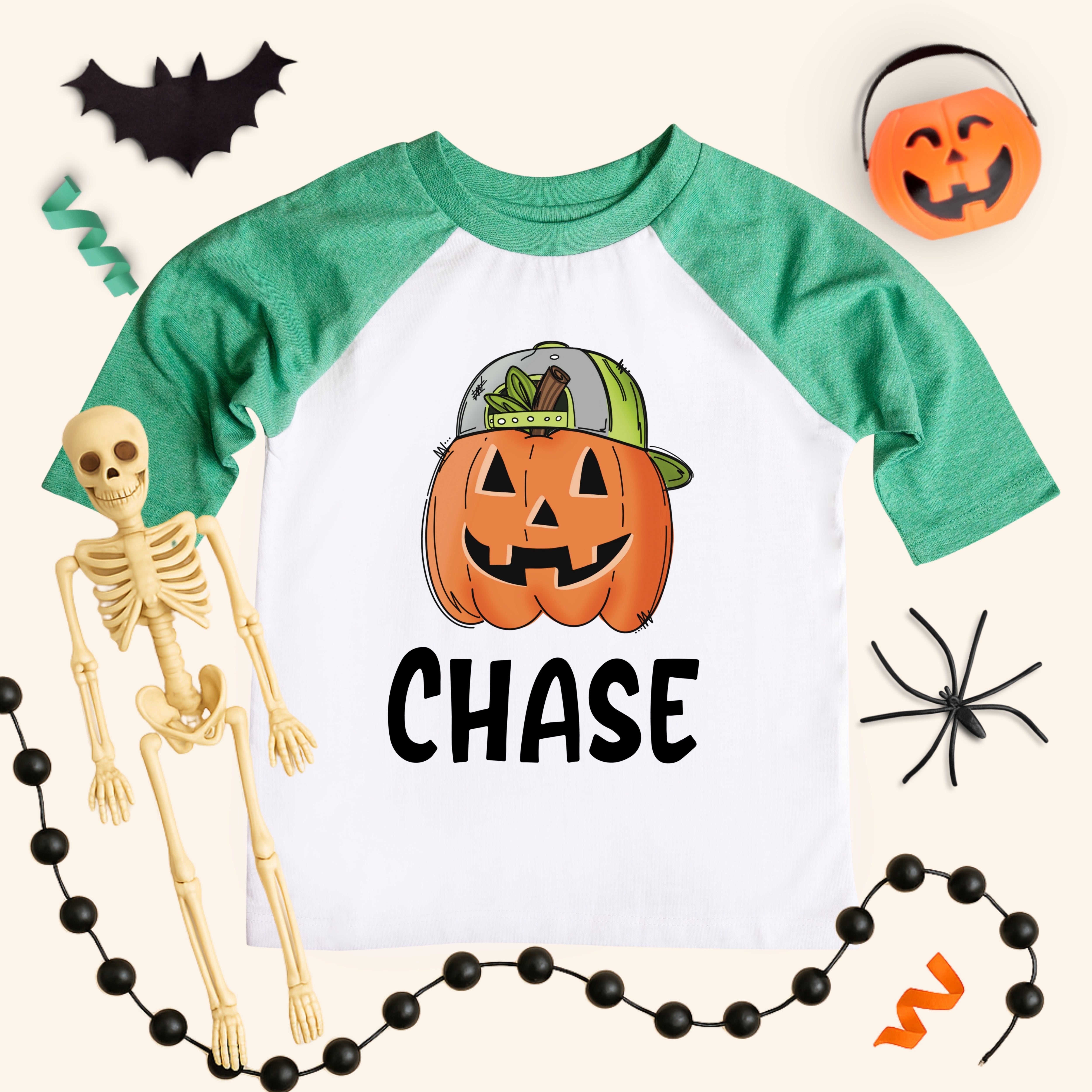 Personalized boys pumpkin shirt on a kids 3/4-sleeve raglan with heather green sleeves and white body. Jack-o’-lantern wearing a cap above the name “CHASE.” Styled flat lay with bat cutout, pumpkin pail, lollipop, spider, skeleton, and black bead garland on a cream background. Kids Halloween name tee, sizes 6M–Youth Large.