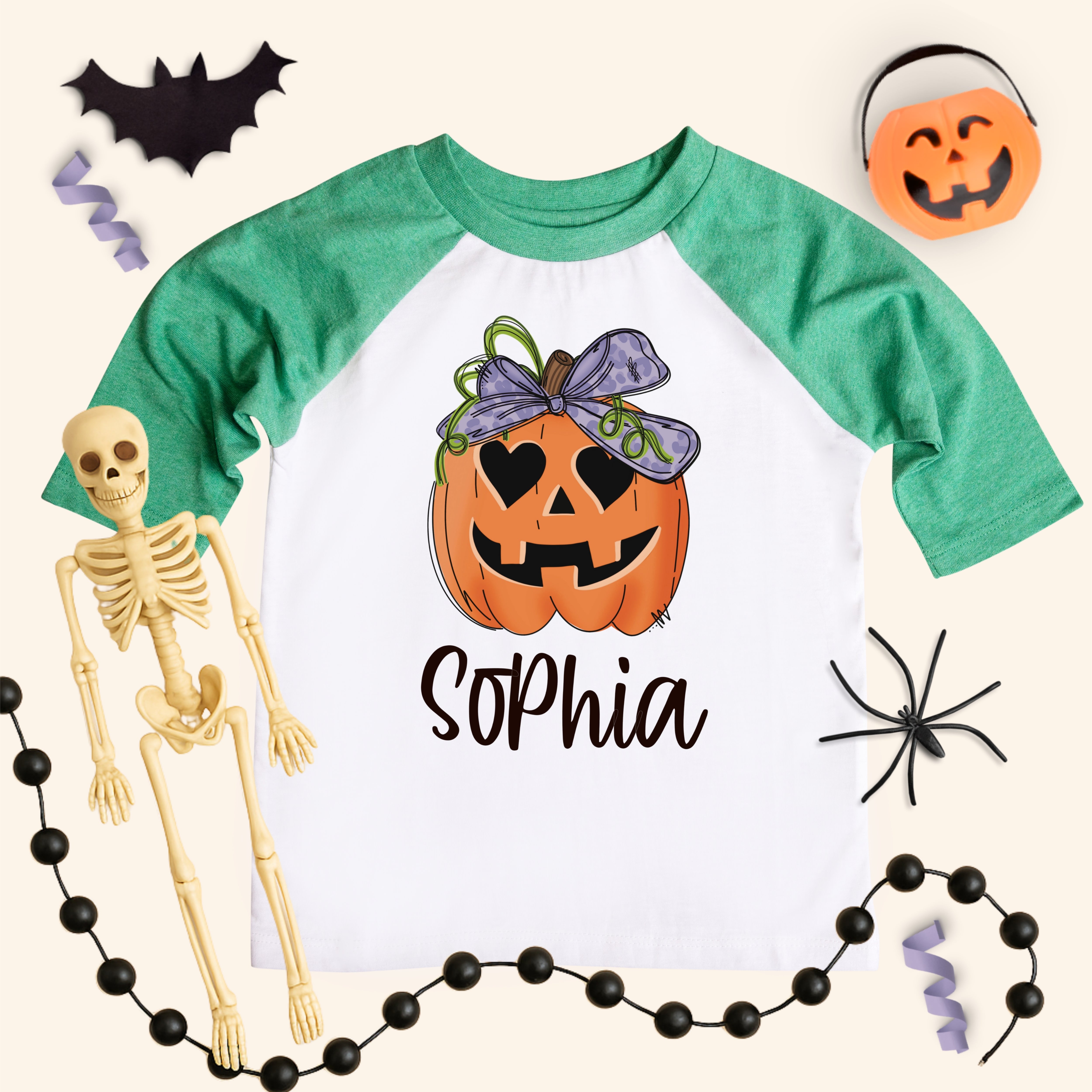 Personalized girls pumpkin shirt on a kids 3/4-sleeve raglan with heather green sleeves and white body. Jack-o’-lantern with a big bow above the name “Sophia.” Flat-lay styling with bat cutout, pumpkin bucket, spider, skeleton, bead garland, and streamers on a cream background. Kids Halloween name tee, sizes 6M–Youth Large.
