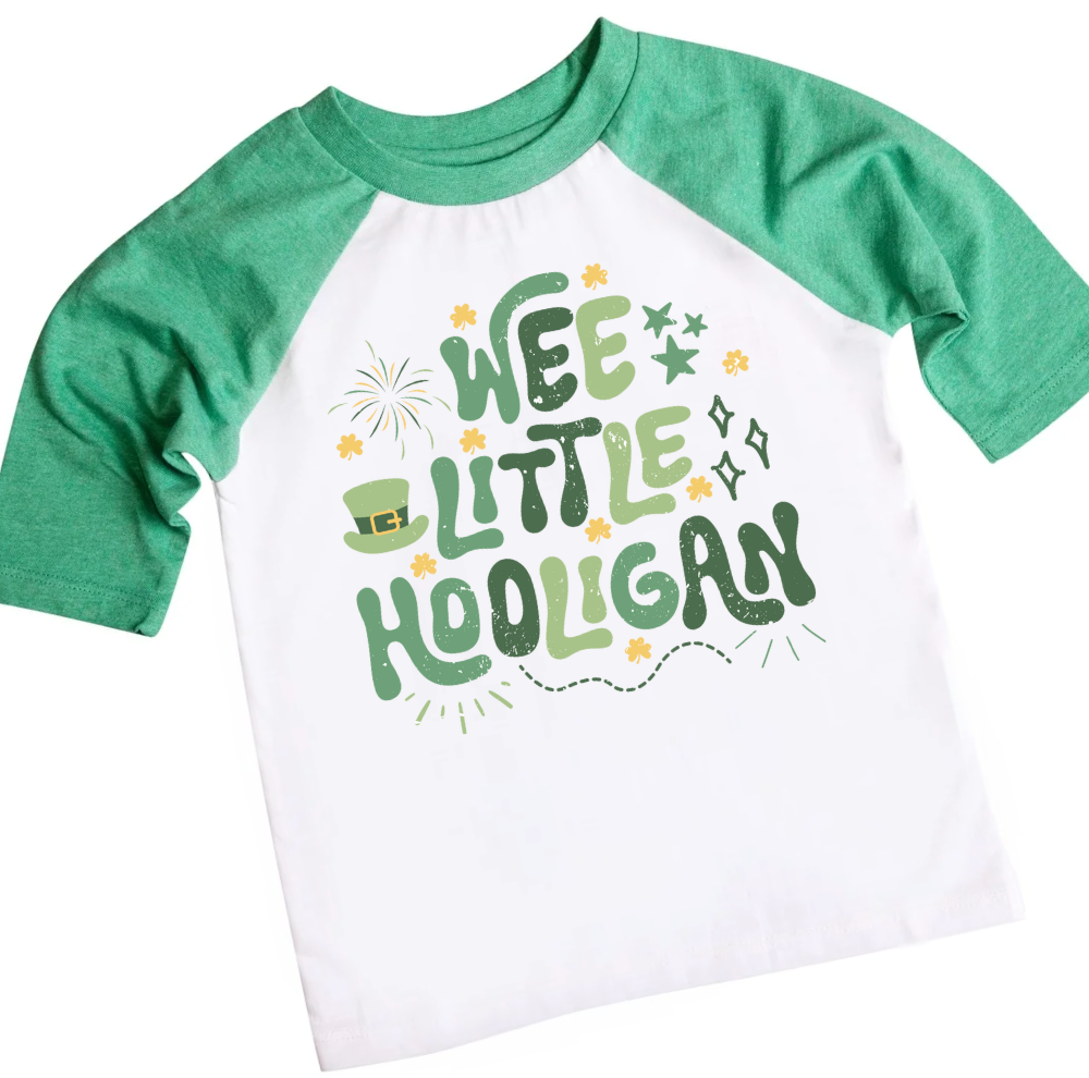 A Wee Little Hooligan Shirt featuring a playful St. Patrick’s Day design with green and gold typography, shamrocks, stars, and a leprechaun hat. The white and blue raglan tee is paired with denim jeans and festive accessories, including gold coins, shamrock beads, and four-leaf clovers, creating a fun and lucky holiday outfit for kids.